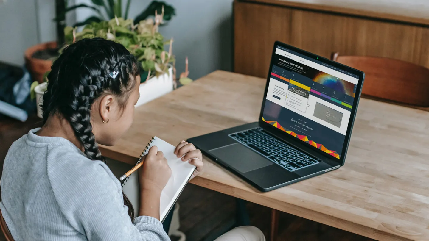 Photo of young girl looking at laptop and taking notes on a notepad